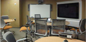 Large video conferencing room with smart board and television/phones for conferencing. Round wooden tables throughout the room